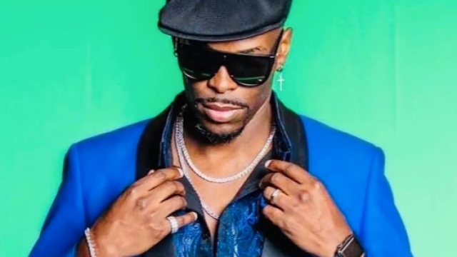 A person wearing a blue blazer, black shirt, black hat, and sunglasses stands against a green background, adjusting their collar with both hands.