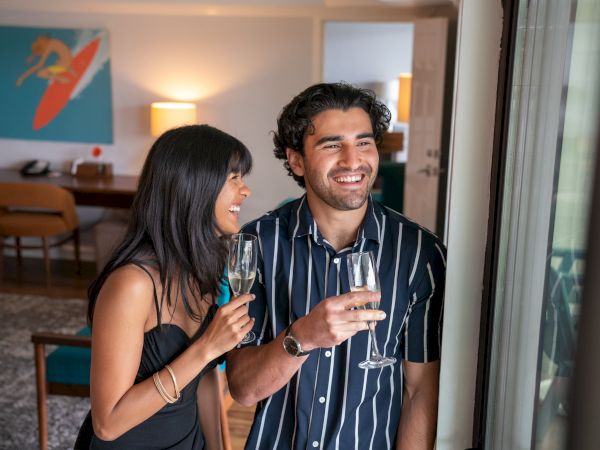 A happy couple stands inside a well-decorated room, holding champagne glasses and smiling while looking out of a window.