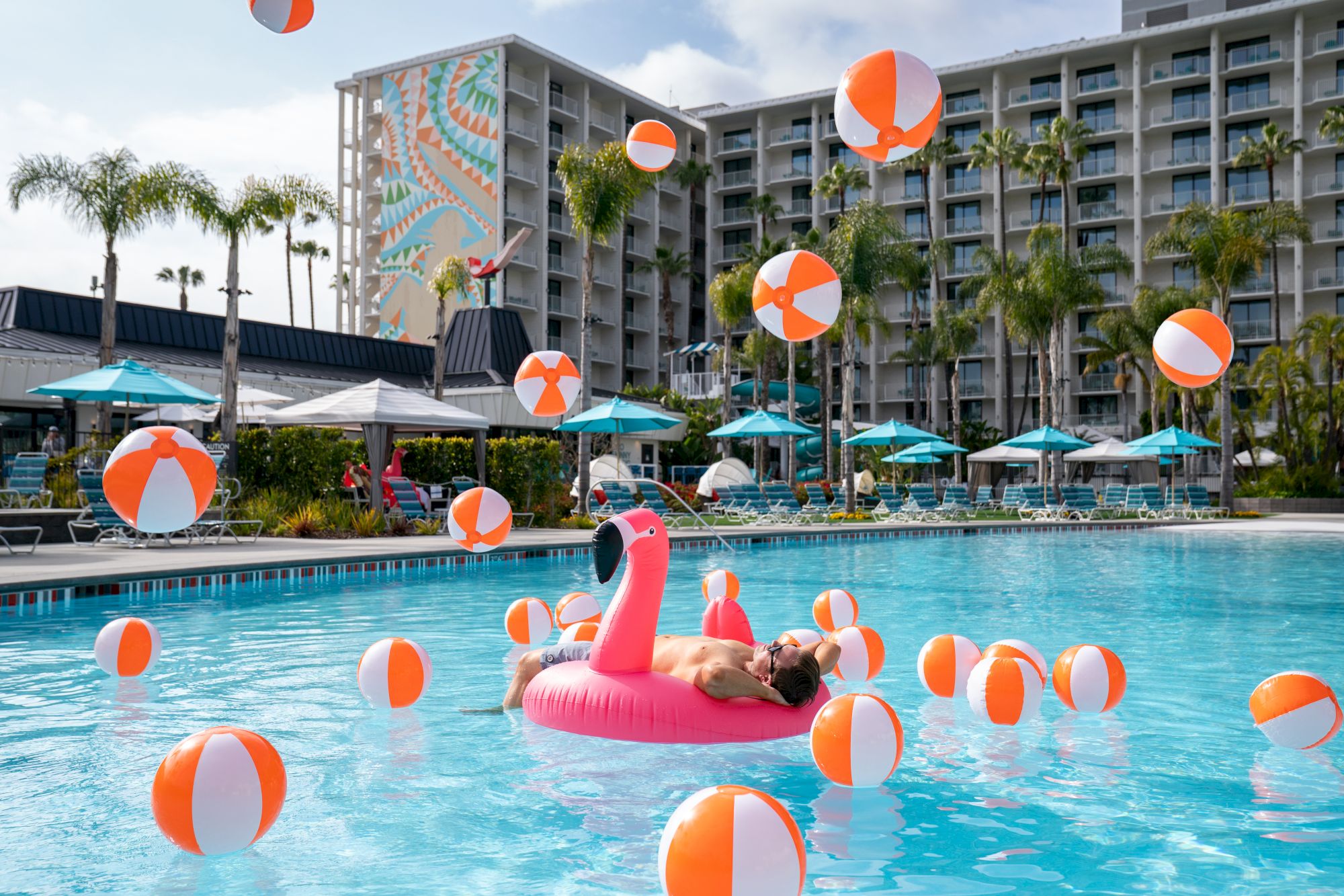 A man lounges on a flamingo float in a hotel pool, surrounded by orange-and-white inflatable beach balls, laughing.