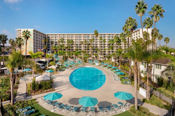 The image shows a large hotel complex with a circular swimming pool, surrounded by sun loungers, umbrellas, and tall palm trees.