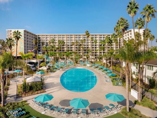 The image shows a large hotel complex with a circular swimming pool, surrounded by sun loungers, umbrellas, and tall palm trees.