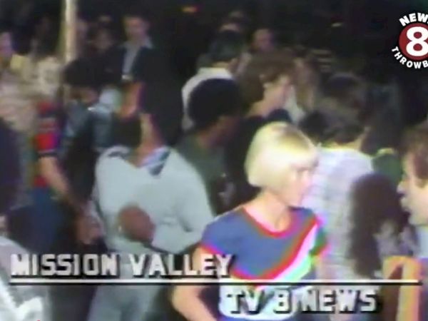 Blurry TV news scene of a crowd at an arena, with a person in a blue-and-orange top in view.