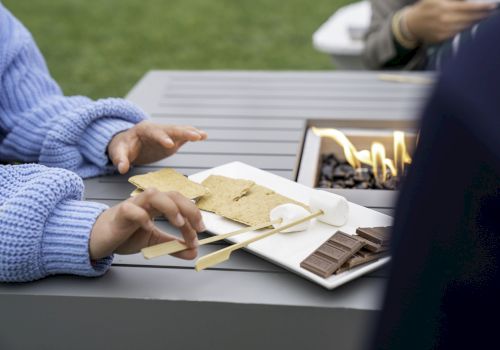 A person in a blue sweater holds skewers with marshmallows near a fire pit, preparing to make s'mores with graham crackers and chocolate on a plate.