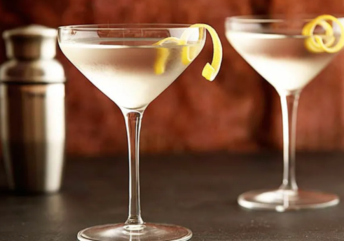 Two martini glasses with lemon twists, alongside a cocktail shaker in the background, on a dark surface with a blurred brown backdrop.