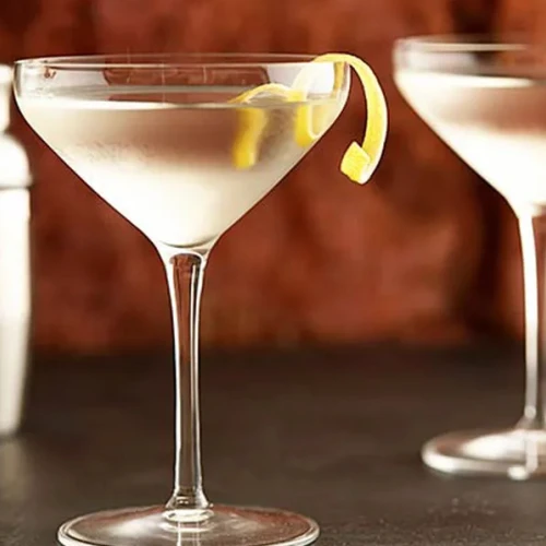 Two martini glasses with lemon twists, alongside a cocktail shaker in the background, on a dark surface with a blurred brown backdrop.