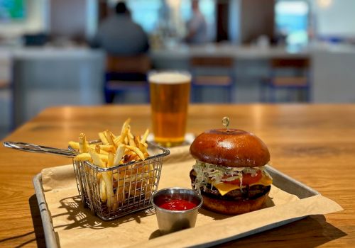 A burger, fries, and a glass of beer on a table in a restaurant; background shows patrons seated at a bar with televisions on the wall.