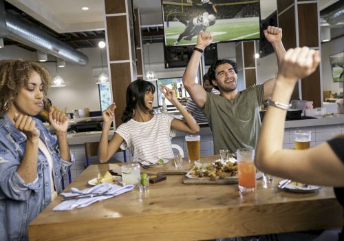 A group of friends celebrates at a table with drinks and food in a bar or restaurant, with a football game playing on the TV.