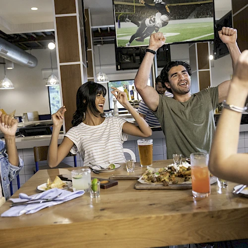 A group of people celebrating at a table with drinks and food, watching a sports game on TV.