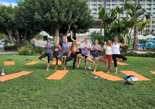 A group of people practicing yoga outdoors on a sunny day with mats on the grass, surrounded by palm trees and buildings in the background.