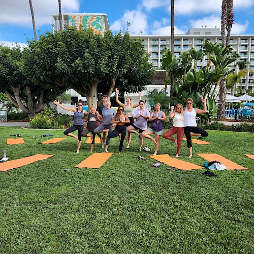 A group of people practicing yoga outdoors on a sunny day with mats on the grass, surrounded by palm trees and buildings in the background.