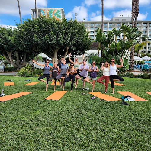 A group of people practicing yoga outdoors on a sunny day with mats on the grass, surrounded by palm trees and buildings in the background.