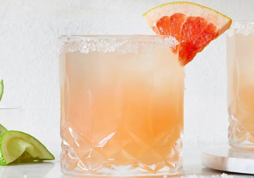 A frosty pink drink in a crystal glass is garnished with a salted rim and a grapefruit wedge; lime slices are visible in the background.