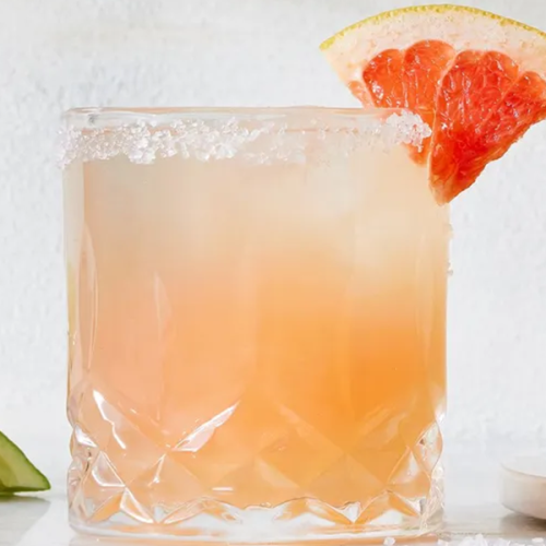 A frosty pink drink in a crystal glass is garnished with a salted rim and a grapefruit wedge; lime slices are visible in the background.