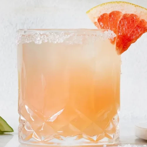 A frosty pink drink in a crystal glass is garnished with a salted rim and a grapefruit wedge; lime slices are visible in the background.