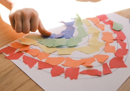 A hand is arranging colorful torn paper pieces into a rainbow pattern on a sheet of white paper, likely part of an art or craft activity.