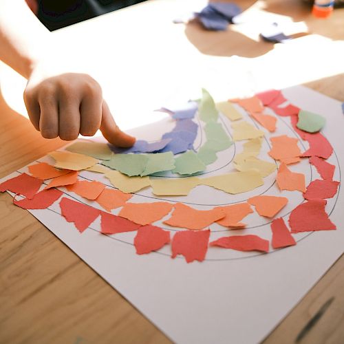A hand is arranging colorful torn paper pieces into a rainbow pattern on a sheet of white paper, likely part of an art or craft activity.