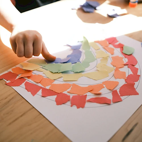 A hand is arranging colorful torn paper pieces into a rainbow pattern on a sheet of white paper, likely part of an art or craft activity.