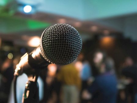A microphone is in focus with a blurred background of people in an indoor setting, suggesting a performance or event.