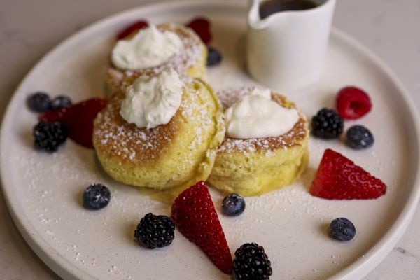 The image shows a dessert plate with three fluffy pancakes topped with cream, surrounded by berries, and served with a small jug of syrup.
