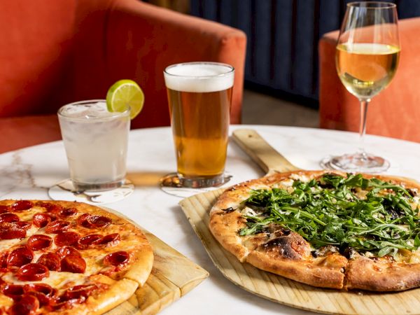 Two pizzas and two drinks&mdash;beer glasses and a lime-topped soda&mdash;sit on a table by the window.