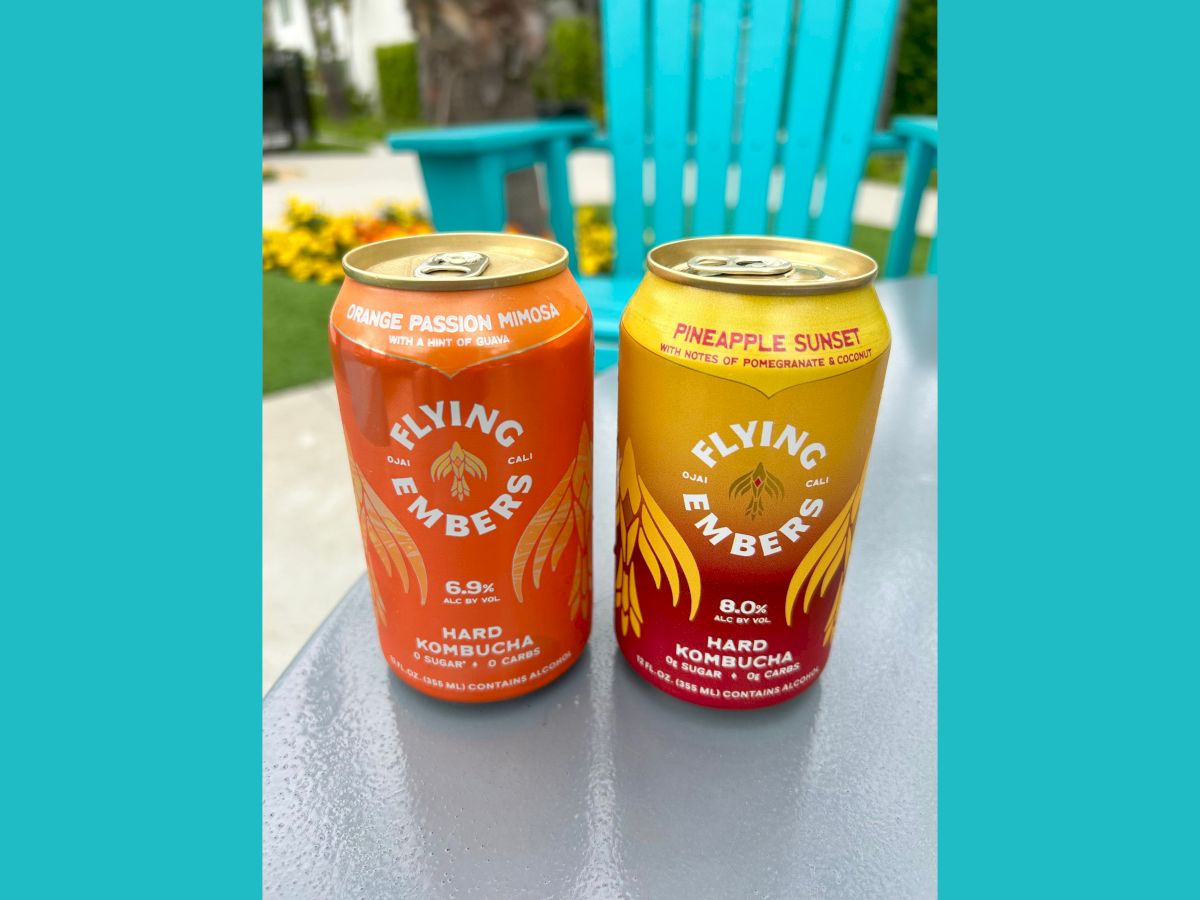 This image shows two cans of Flying Embers Hard Kombucha in different flavors, Orange Passion Mimosa and Pineapple Sunset, on an outdoor table.