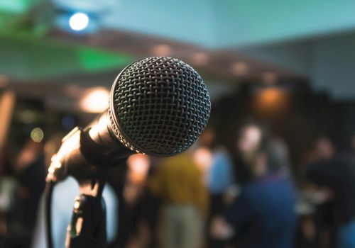 The image features a close-up of a microphone with a blurred background, possibly at an event or gathering with people mingling.