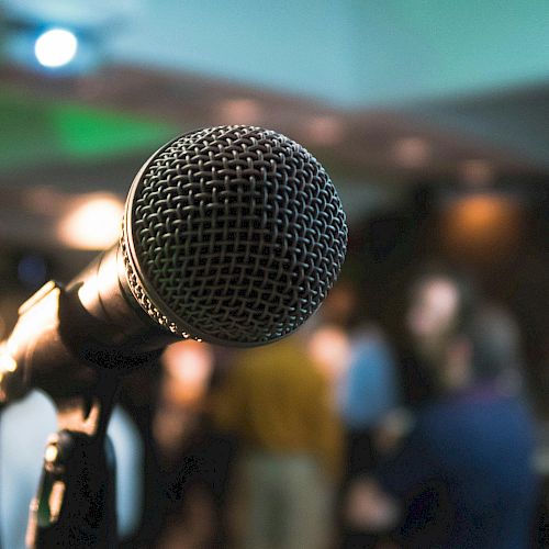 The image features a close-up of a microphone with a blurred background, possibly at an event or gathering with people mingling.