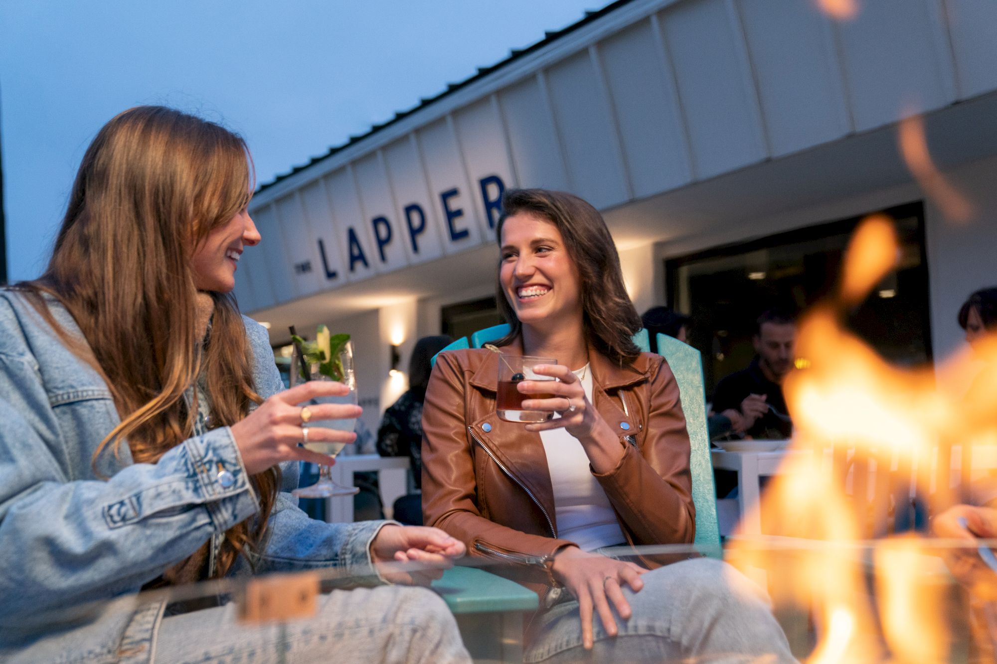 Two women are enjoying drinks and laughing by a fire pit at an outdoor venue with a sign that reads "LAPPER" in the background.