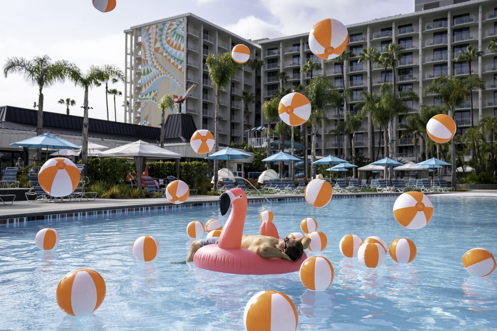 A person relaxes on a flamingo float in a pool surrounded by numerous beach balls, with a hotel and palm trees in the background that's located close to San Diego Universities and colleges including San Diego State University, University of San Diego, University of California San Diego and Point Loma Nazarene University.