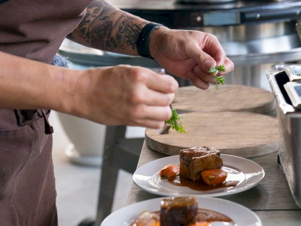 A chef is plating a dish with meat and vegetables, adding garnish to two plates in a kitchen setting.