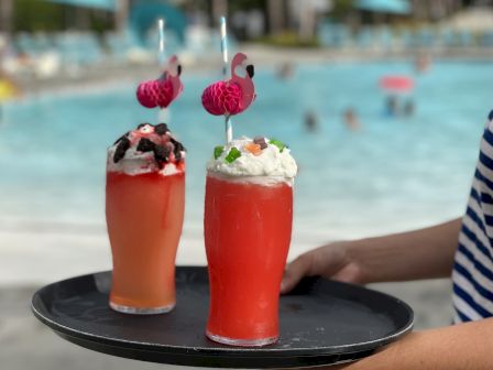 Two red poolside drinks with pink umbrella toppers sit on a tray while a blurred swimming pool stretches behind them.