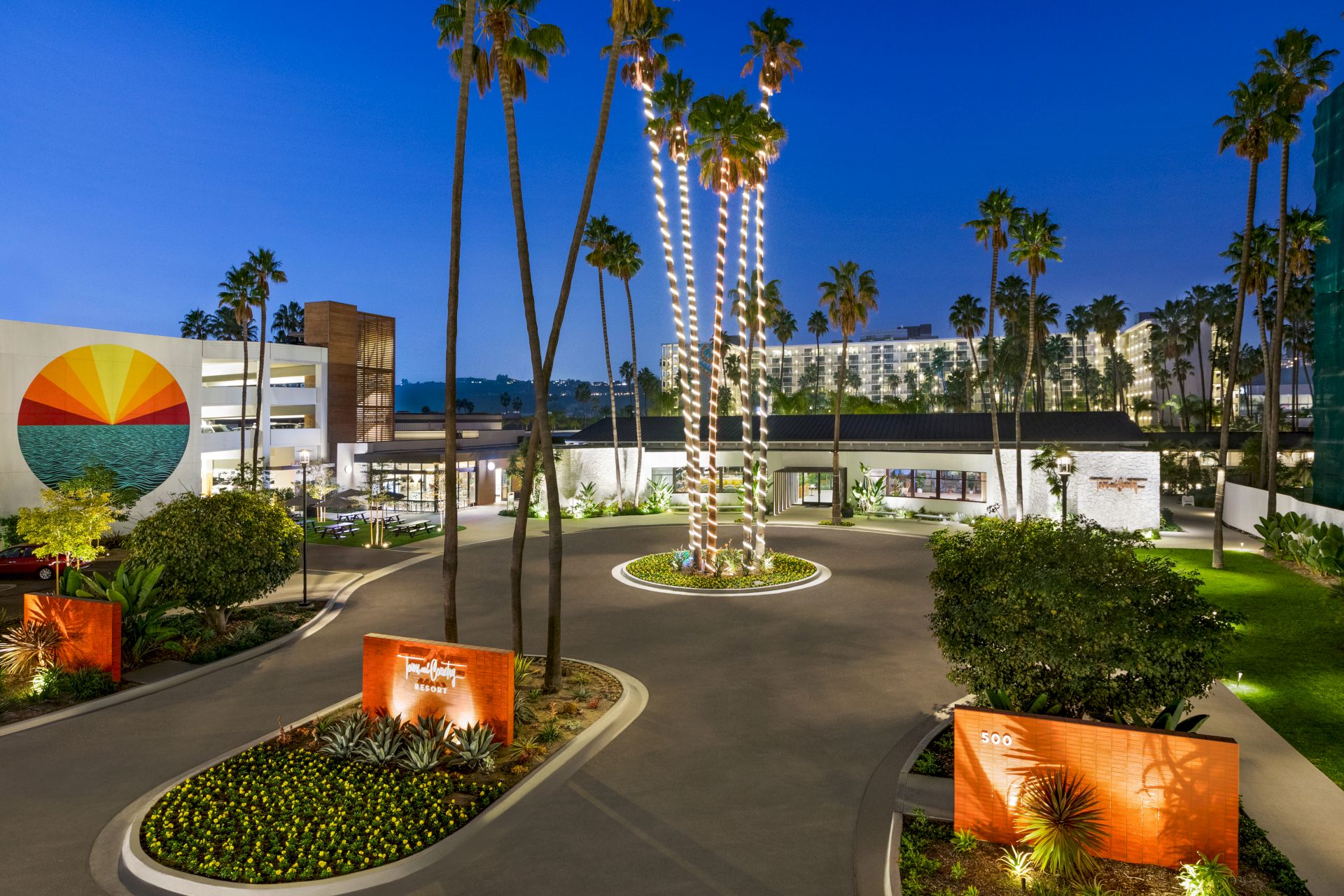 An illuminated courtyard with tall palm trees, landscaped greenery, and a building adorned with a sun mural.