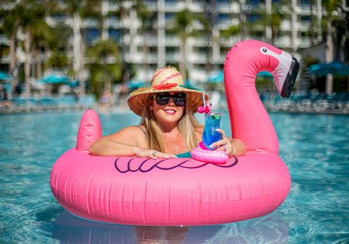A woman in a sunhat and sunglasses relaxes in a flamingo float in a pool, holding a cocktail with a small flamingo decoration, with buildings behind.