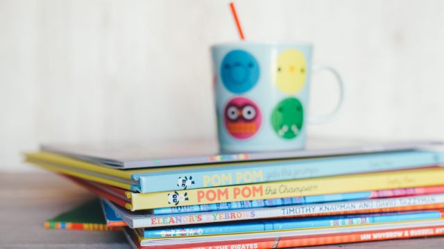 A stack of children's books with a colorful mug on top, featuring playful faces and a red straw, placed on a wooden surface.