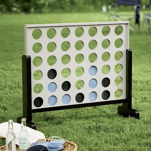 The image shows an outdoor Connect Four game set on grass, accompanied by drinks and a basket with plates.