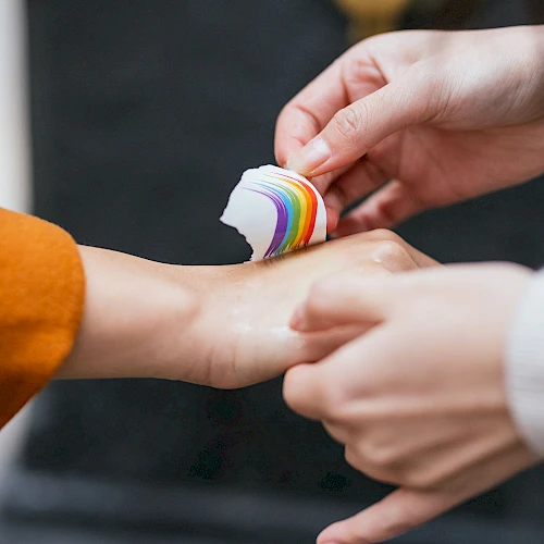 Two hands are seen, one person is applying a rainbow-colored bandage to the wrist of the other person&rsquo;s hand, which is slightly outstretched.