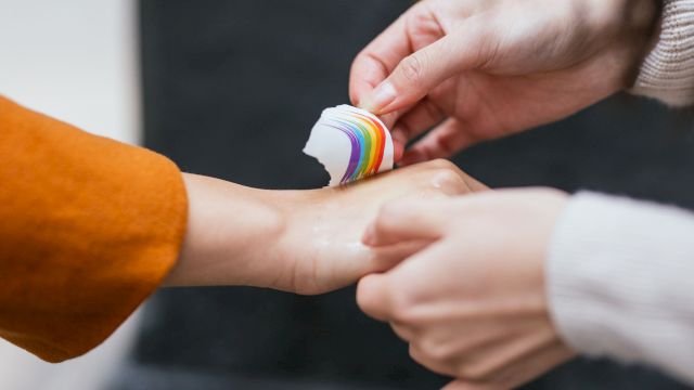 Two hands are seen, one person is applying a rainbow-colored bandage to the wrist of the other person’s hand, which is slightly outstretched.