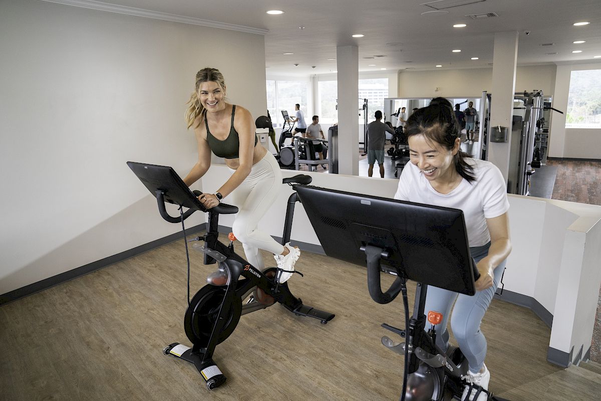 Two women are riding stationary bikes in a gym. They appear to be smiling as they exercise. Other gym equipment can be seen in the background.