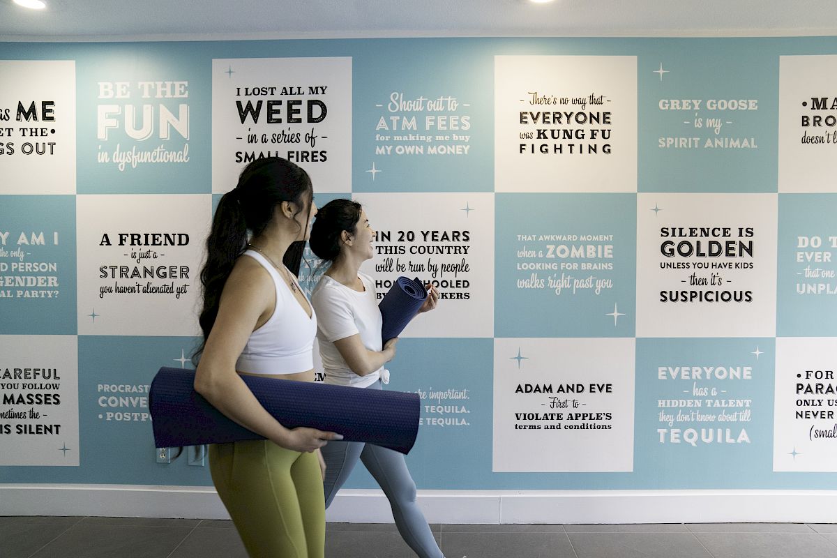 Two people are walking with yoga mats in front of a wall covered with humorous and motivational quotes.