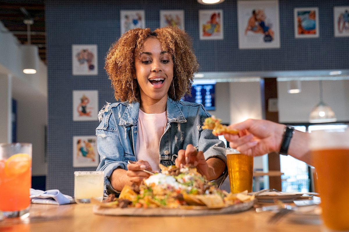 A woman happily reaches for food at a table with various drinks, eagerly anticipating a delicious meal in a casual dining setting.