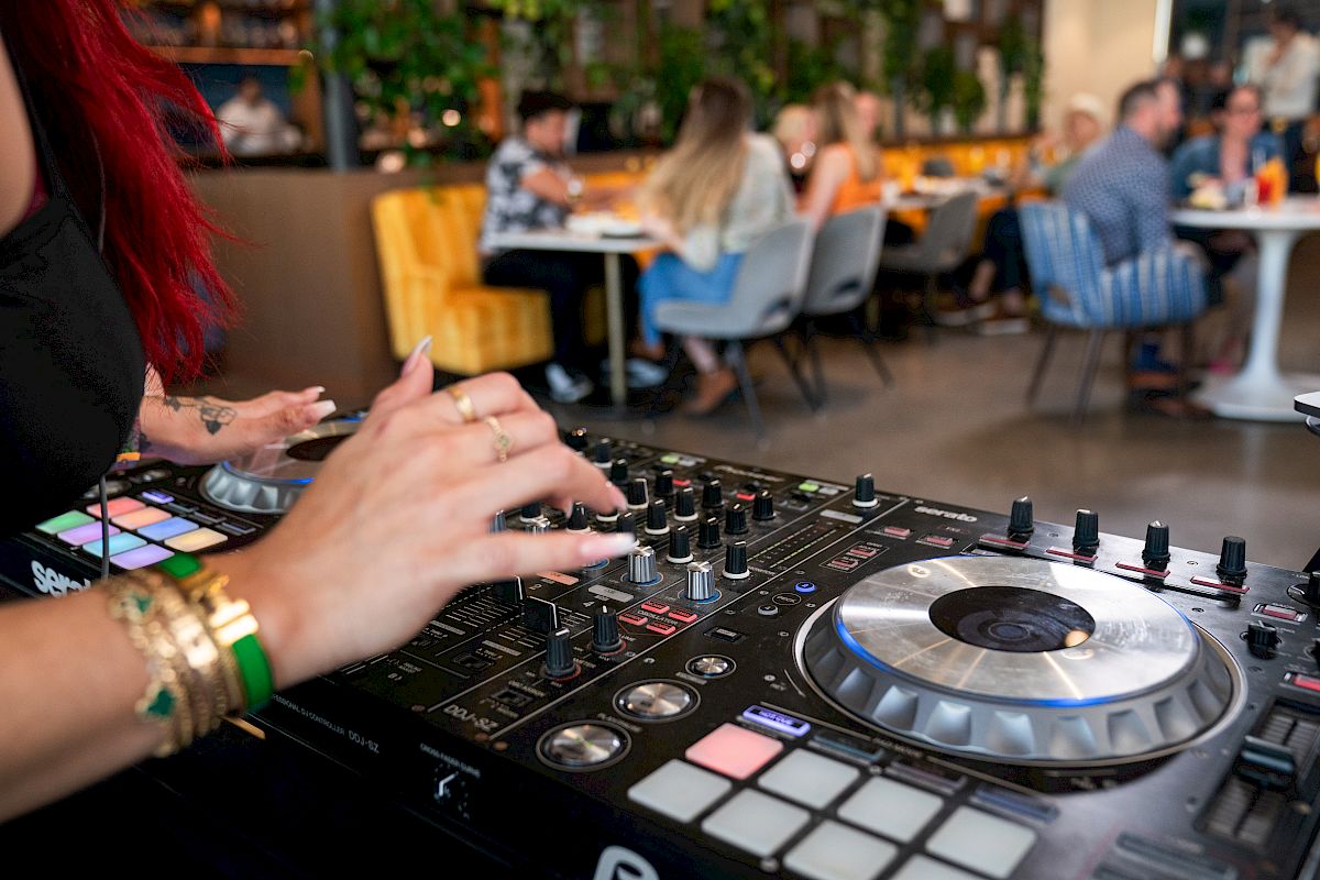 A DJ is performing while people sit and socialize at tables in a modern indoor venue with greenery and stylish furnishings.