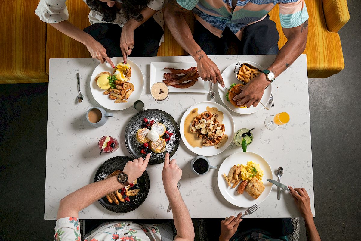Four people are eating a meal at a restaurant, with dishes including pancakes, bacon, eggs, and coffee on a white marble table.
