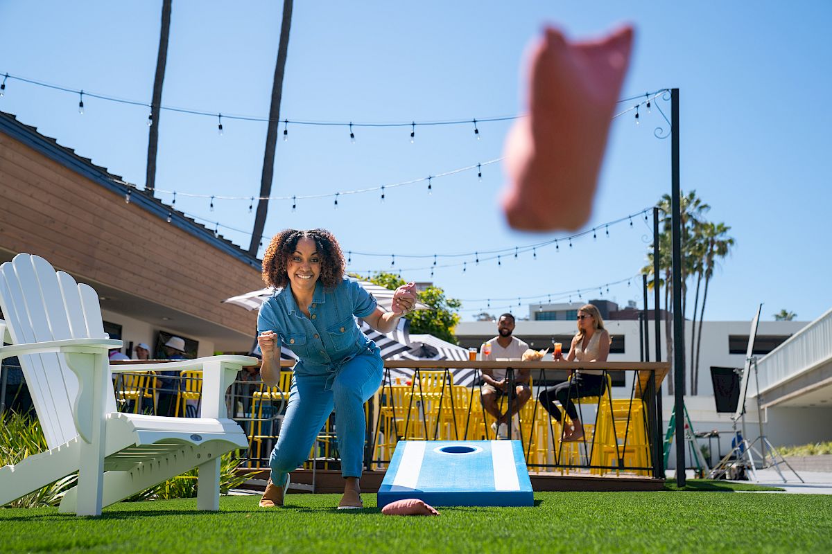 A person is throwing a beanbag towards a cornhole board outside while others sit and chat nearby on a sunny day.
