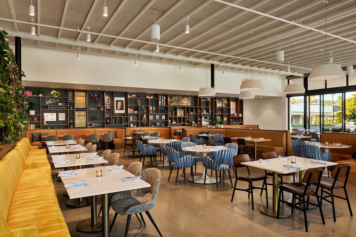 The image shows a modern restaurant interior with tables, chairs, a yellow couch, book shelves, and large windows letting in natural light.
