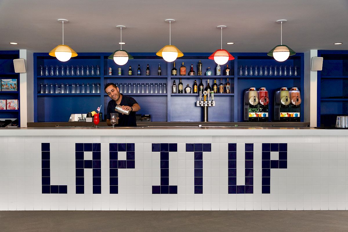 A bartender preparing drinks behind a counter with the words "LAP IT UP" on the front. Shelves of bottles and drink machines are in the background.