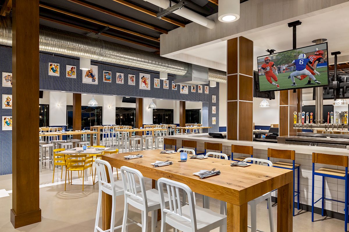 A modern restaurant with wooden tables and white and yellow chairs, decorated with sports images, a bar area, and a TV showing a football game.