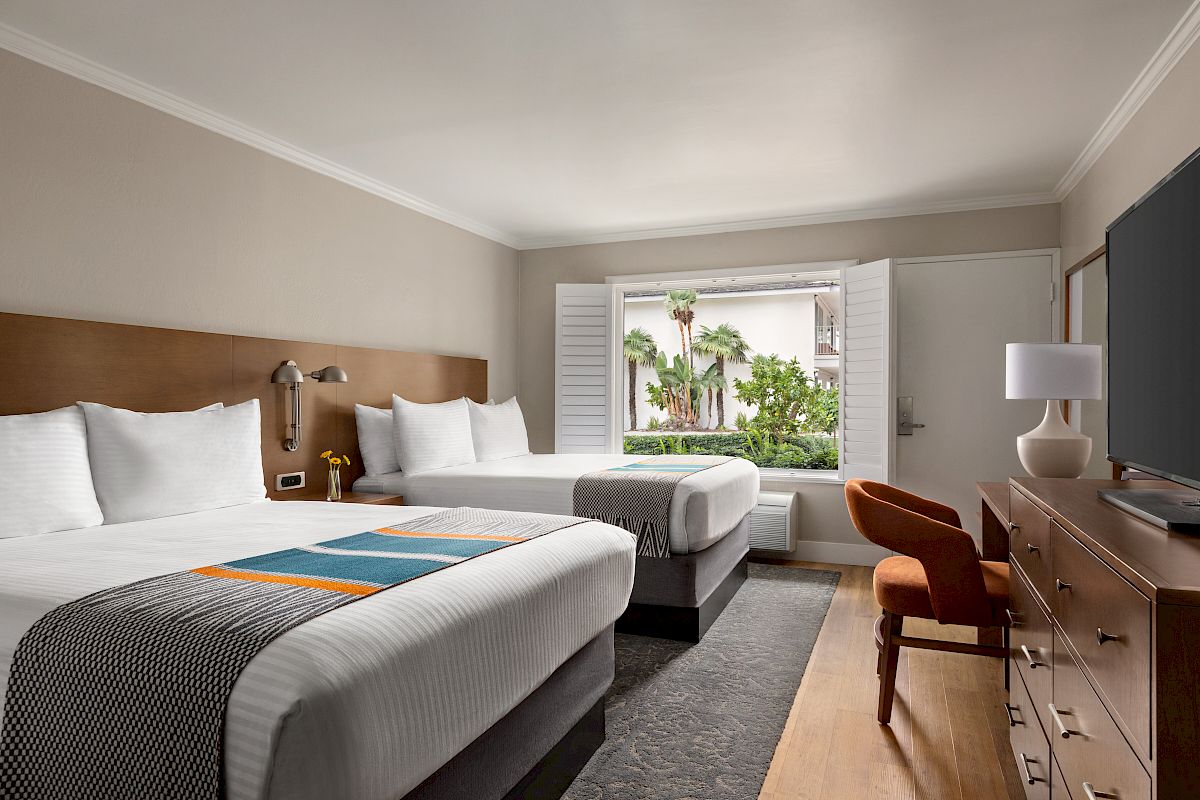 This image shows a hotel room with two beds, a window with open shutters, a TV, a dresser, and a chair, all in a modern, clean design.
