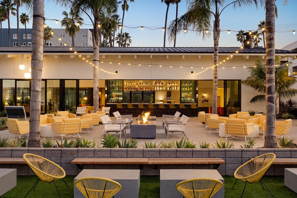 This image depicts an outdoor lounge area with yellow chairs, palm trees, string lights, and a central fire pit in front of a modern bar building.
