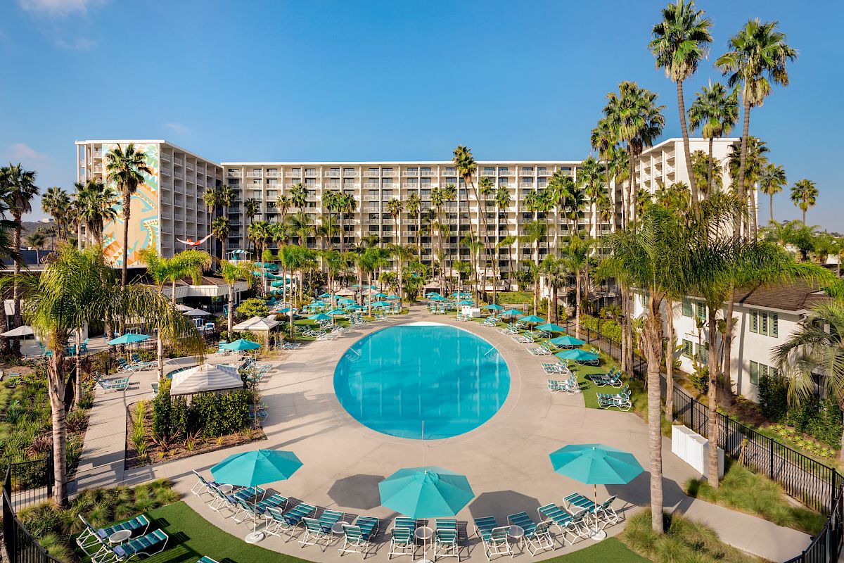 The image shows a large hotel with a tall building, numerous palm trees, and a central round swimming pool surrounded by lounge chairs and umbrellas.
