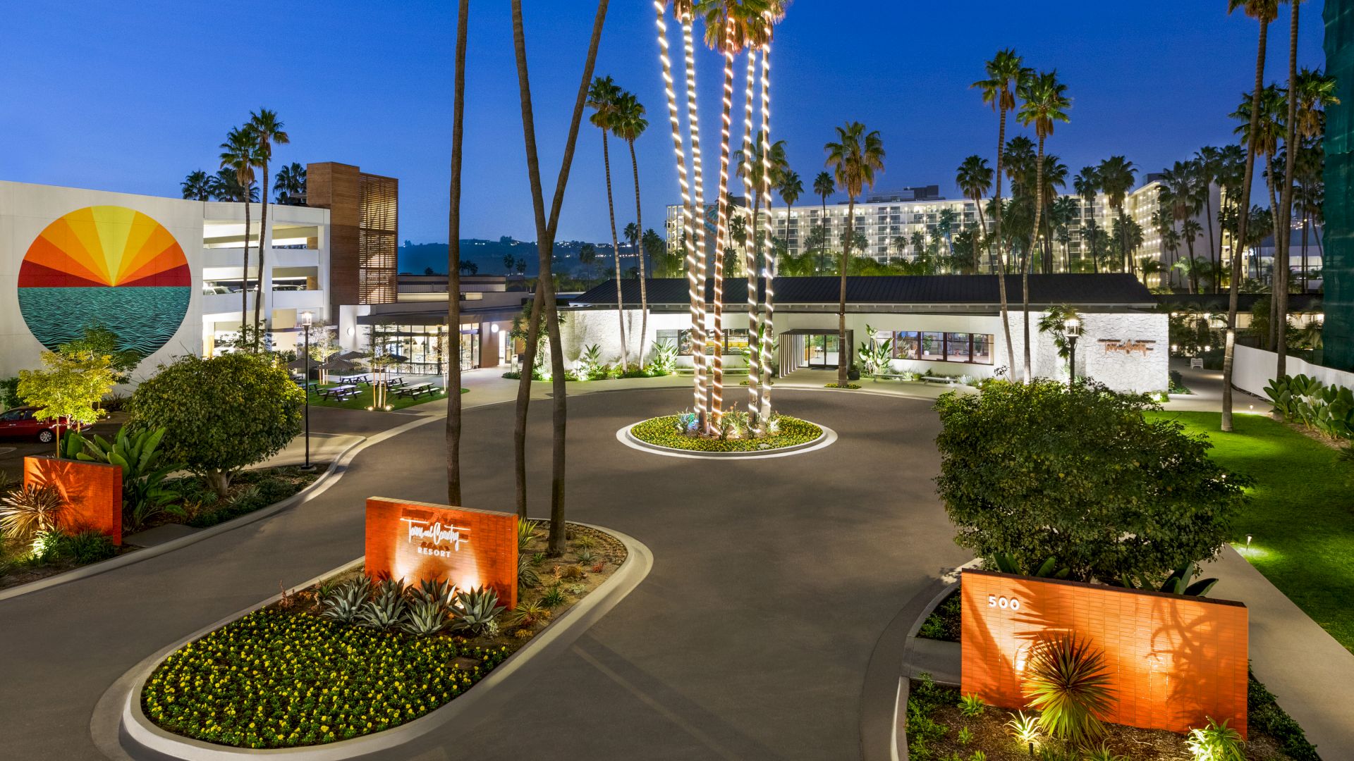 The image shows a hotel entrance with palm trees, landscaped greenery, and modern architecture, featuring a mural of a sunset on a building. Town and Country Resort is a hotel that has trolley access to Snapdragon Stadium, Viejas Arena, Petco Park, and the Rady Shell.